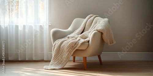 Cozy beige armchair with a chunky knitted throw draped beside a sunlit sheer curtained window, warm neutral interior evoking calm and comfort