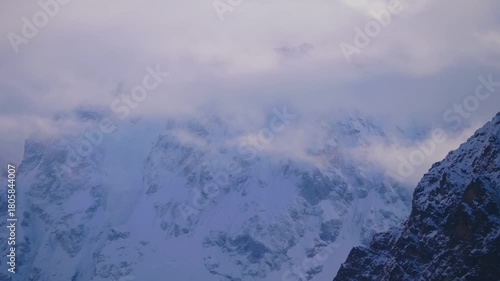 A view of the snow covered peaks of the North Caucasus mountains in Russia, partially shrouded in clouds. The scene captures the vast, rugged beauty of the mountain range under a dramatic sky, evoking