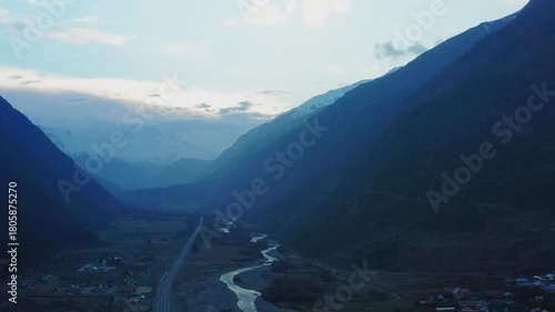 An aerial shot captures the majestic beauty of the North Caucasus mountains in Russia. A serene river snakes through a vast valley, paralleled by a winding road. Small, scattered villages are visible,