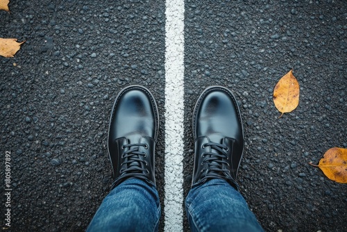 top-down view of black leather boots in blue jeans standing on either side of a white dividing line on asphalt with fallen autumn leaves, contemplative mood