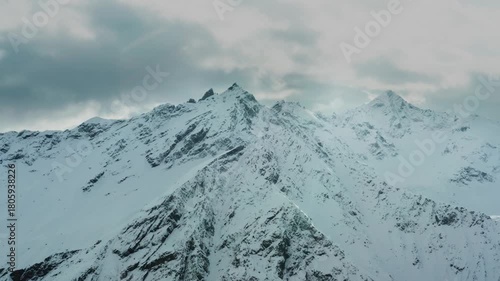 An aerial shot of the stunning, snow covered mountains of the North Caucasus region in Russia. The footage captures the rugged beauty of the mountain range under a dramatic, cloudy sky, highlighting t