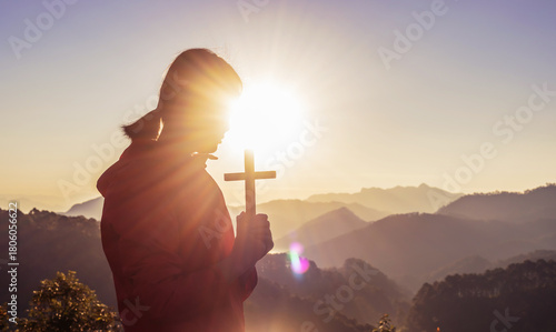 Girl praying and holding Christian cross for worshipping God on mountain at sunrise background. Christian, Christianity, Religion copy space background. Easter Sunday concept: