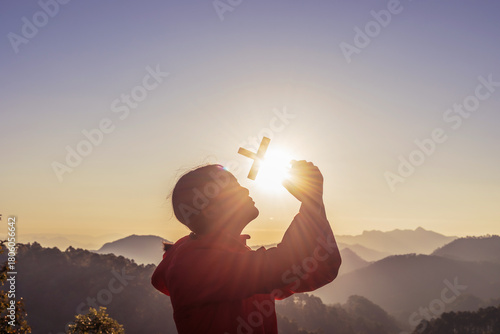 Girl praying and holding Christian cross for worshipping God on mountain at sunrise background. Christian, Christianity, Religion copy space background. Easter Sunday concept: