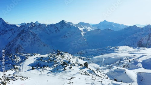 aerial view of a snow covered mountain range in the North Caucasus, Russia. The footage captures the vast, pristine winter landscape under a clear blue sky, with ski slopes and a small cluster of buil