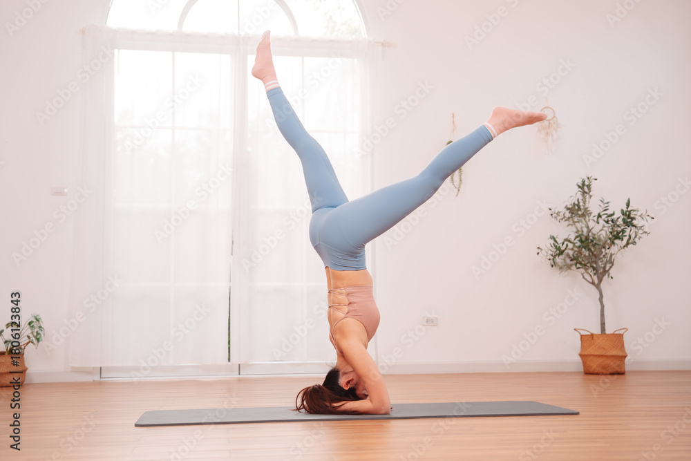 Obraz premium Young Asian woman practicing advanced yoga headstand pose indoors on a mat. Concept of balance, strength, focus, flexibility, and body control in a calm and minimalist wellness environment.