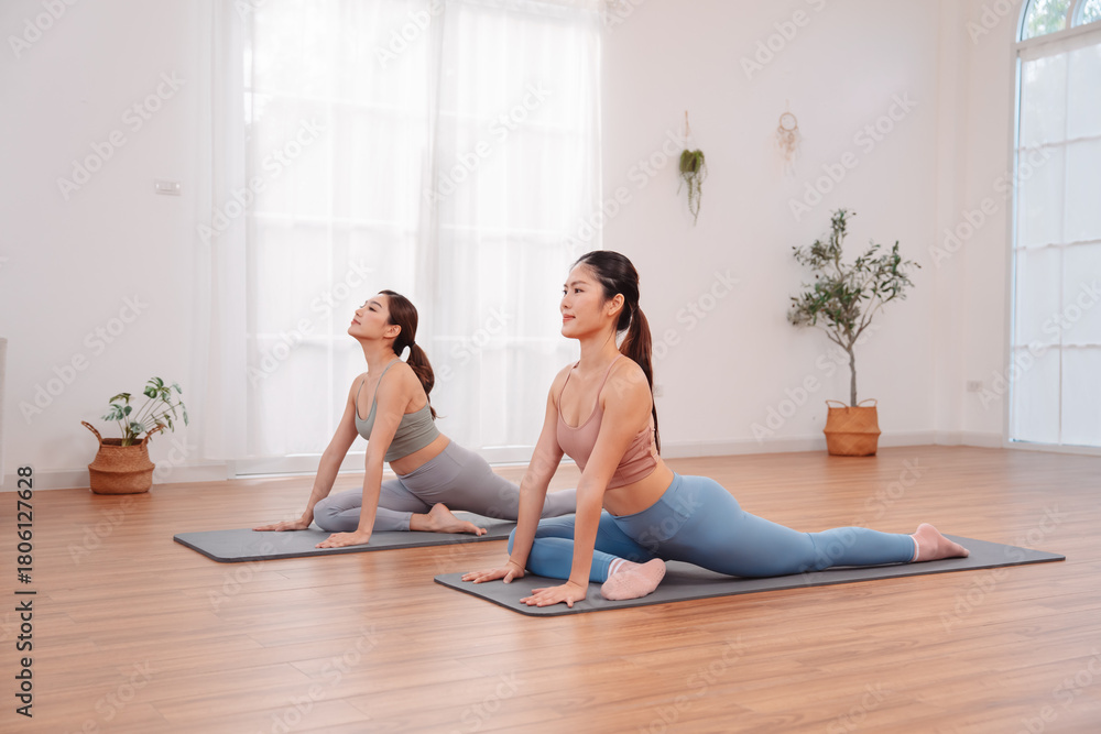Obraz premium Two Asian women practicing yoga cobra pose together on mats. indoor space, flexibility, healthy lifestyle, relaxation, and body alignment through stretching exercise.