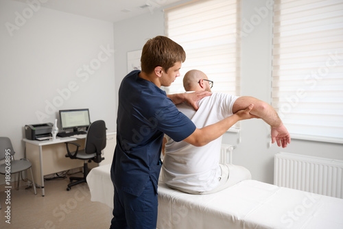 Chiropractor adjusting male patient’s shoulder in clinic. Physiotherapist performing manual therapy to relieve pain, improve mobility, support rehabilitation after injury or musculoskeletal disorder.