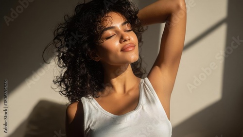 Curly-haired woman in a white tank top soaking up warm sunlight with eyes closed and one arm raised. Concept Curly-haired woman, White tank top, Sunlit warmth, Eyes closed, Arm raised in golden light