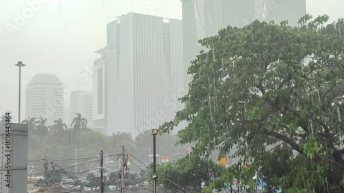 Rain falls heavily on the city streets during a summer downpour, creating a wet and gloomy atmosphere with skyscrapers in the distance.