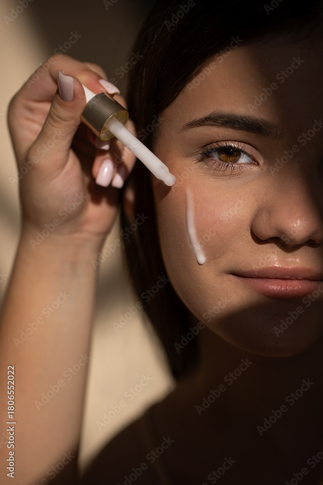 Obraz premium Young teen girl applying skincare serum to her face with a dropper, showcasing a beauty routine in soft lighting, highlighting skin texture and natural beauty