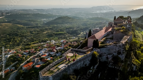 Aerial view of a medieval fortress perched atop a hill, overlooking a quaint village nestled amidst lush greenery, Klis, Croatia.