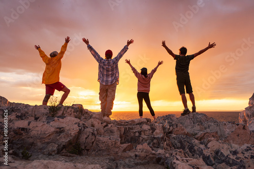 Four happy diverse tourists are having fun and meeting sunset with open arms at lake shore against colorful pink and orange cloudy sky. Active vacations concept