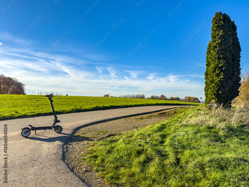 Fototapeta premium Electric scooter parked on a winding country road beside a tall cypress, fresh green grass and wide blue sky. Peaceful countryside scene in Luxembourg.
