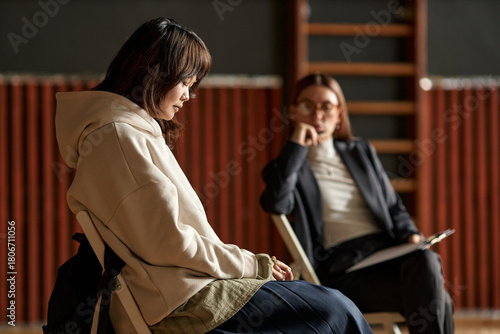 Asian female teenager sitting with head down during psychology session while Caucasian young adult woman psychologist observing and taking notes in educational setting