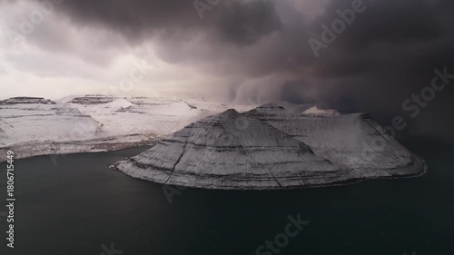 Aerial view of snow-dusted mountains rise from the dark waters, under a heavy, brooding sky, creating a stark contrast of light and shadow, Streymoy, Faroe Islands.
