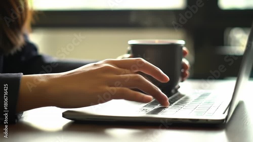 A person uses a laptop, one hand typing, the other holding a mug. The scene is indoors with soft lighting