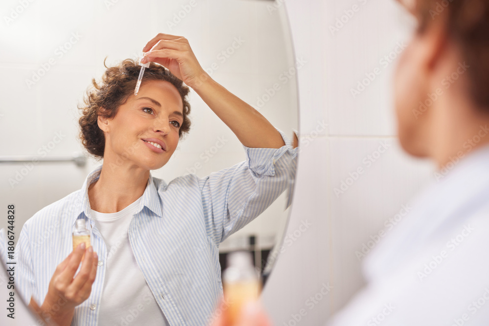 Fototapeta premium A woman smiles as she applies serum to her forehead in front of a mirror. The bright bathroom adds a fresh vibe, showcasing her morning skincare routine.
