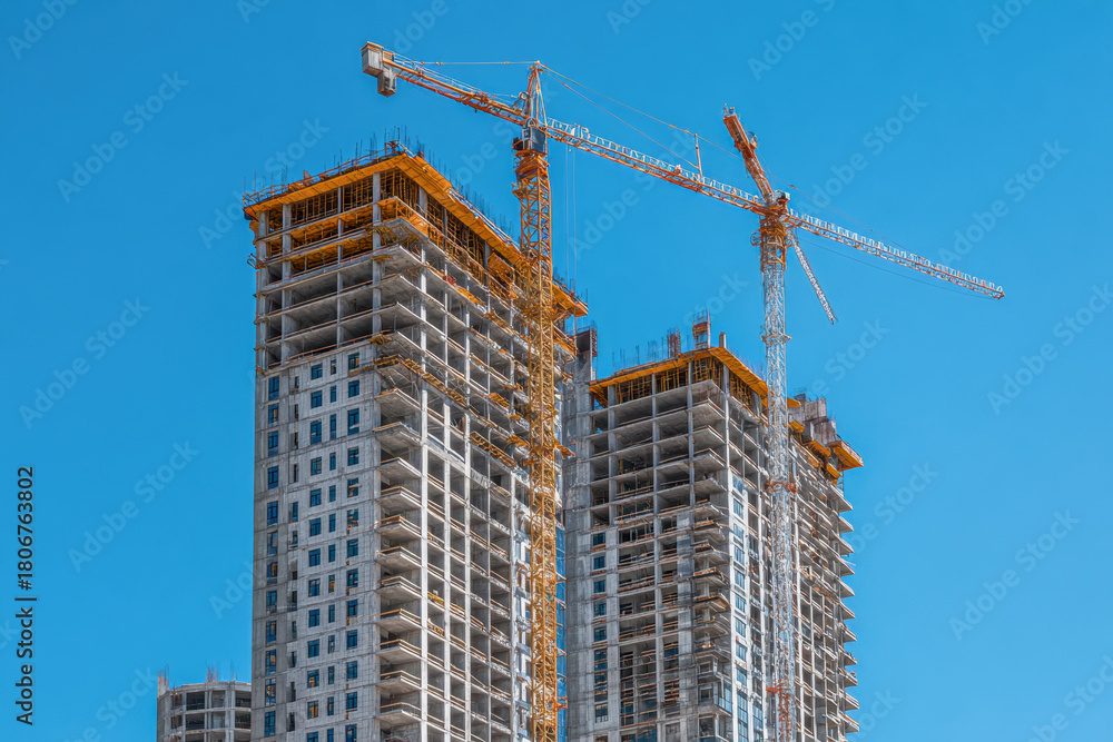 Fototapeta premium Construction of modern high-rise buildings featuring towering cranes against a clear blue sky in an urban development site
