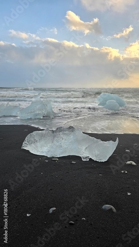 Crystal-clear iceberg fragments shimmer on the black volcanic sand of Diamond Beach, Iceland, as golden sunrise light reflects on waves and ice.