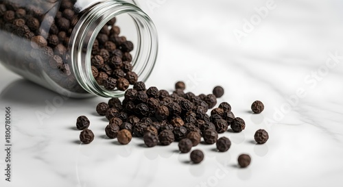 Black peppercorns spilling out of a glass jar onto a white marble surface.