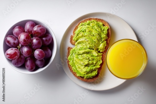 An overhead flat lay of avocado toast , a glass of fresh orange juice, and a small bowl of purple grapes, on a clean white table, 