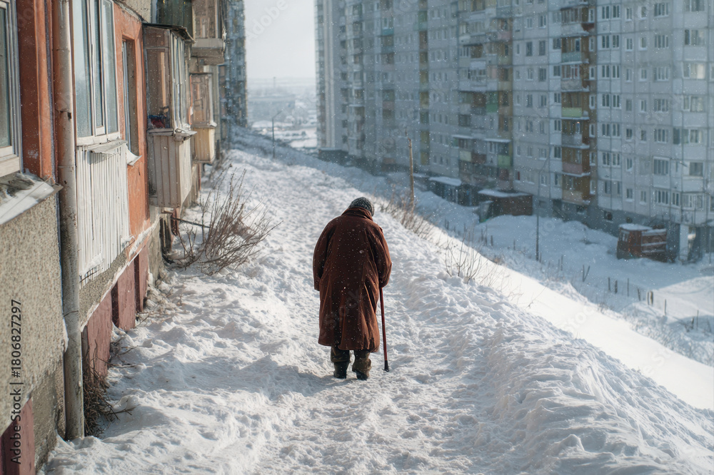 Naklejka premium An elderly woman in a coat walks through the snow leaning on a cane in a winter city