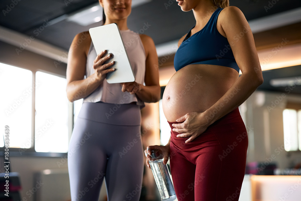 Naklejka premium Close up of pregnant woman and her fitness instructor using touchpad at health club.