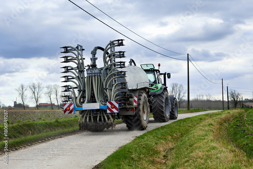 A green tractor pulls a large farm implement on a rural road under a cloudy sky. Power lines stretch across the rustic landscape, showcasing modern agriculture and countryside transport.