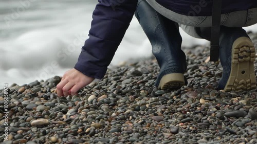 Boots, pebbles, beach. Woman in rubber boots carefully picking up smooth stones near the crashing sea.