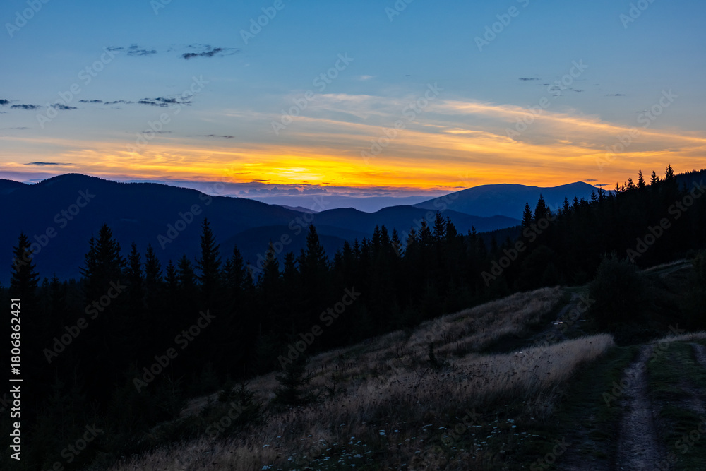 Obraz premium A serene twilight panorama of layered mountains and dark conifer silhouettes beneath a vivid orange and blue sunset, with a dirt path across a grassy hillside inviting wonder and calm reflection.