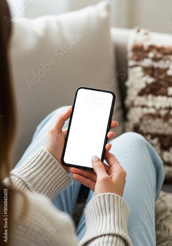 Woman holding a blank smartphone with a white screen while relaxing on a couch in a cozy living room setting perfect for showcasing mobile app designs