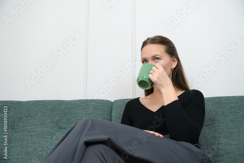 Young woman sitting comfortably on a green sofa, savoring a moment of relaxation while enjoying a warm beverage from a green mug in a cozy home setting