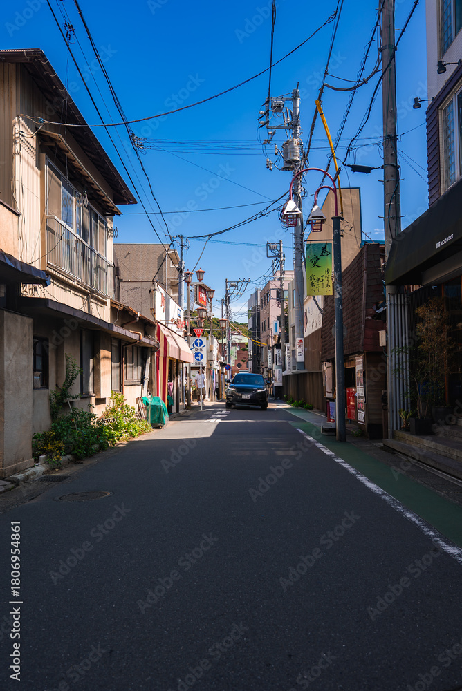 Fototapeta premium A narrow Kamakura street shows wooden and concrete buildings, power lines, a small truck, vending machines, and plants in bright midday sun with crisp shadows.