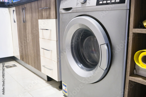 Modern front-loading washing machine integrated into contemporary kitchen with wooden cabinets and tiled floor. Home appliances and interior design in clean, bright household environment