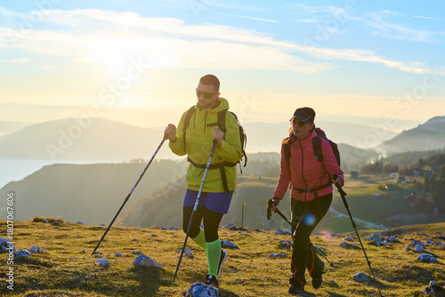 Couple hiking through a grassy mountain trail during sunrise, surrounded by a stunning backdrop of fog in the valleys below. The warm light creates a beautiful atmosphere