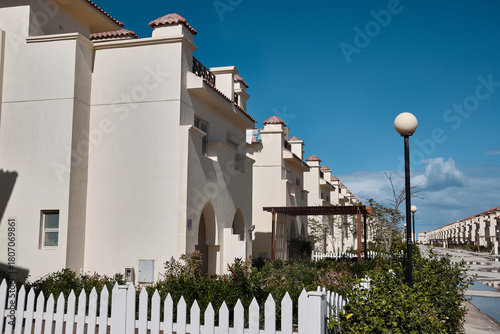 Line of modern white residential houses with red-tiled roofs under clear blue sky. Bright architecture, white fence and sunny weather create calm suburban atmosphere