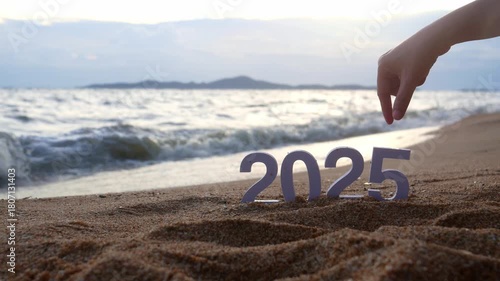 hand picks out the number 5, changing from year 2025 to 2026 for new year sign on summer beach at sunset, with ripple sea wave and beautiful nature at background, 2026 New Year celebration concept