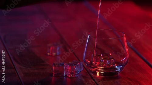Whiskey pours into glass with ice cubes standing on the wooden table on the dark background. Close-up. Nightlife theme. Slow motion.