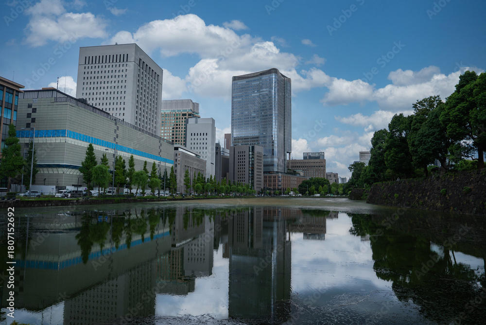 Fototapeta premium Daytime view in central Tokyo shows a canal reflecting glass towers near the Marunouchi district and the Imperial Palace moat, with trees and stone embankment in frame.