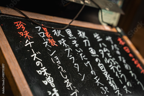 Photography Close up chalk board shows vertical kanji and red headers for specials at a Tokyo izakaya entrance, shallow angle and tight framing highlight wood texture and daily dishes