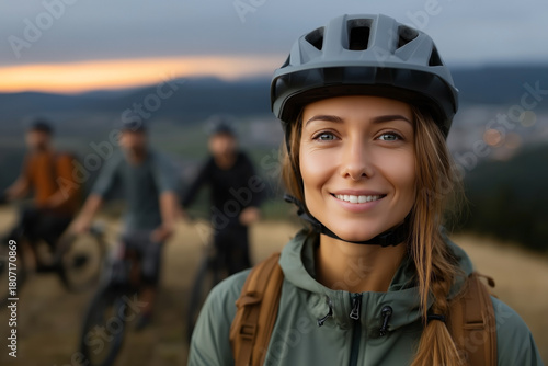 Smiling young woman wearing helmet with friends cycling in background, scenic hills at sunset behind group