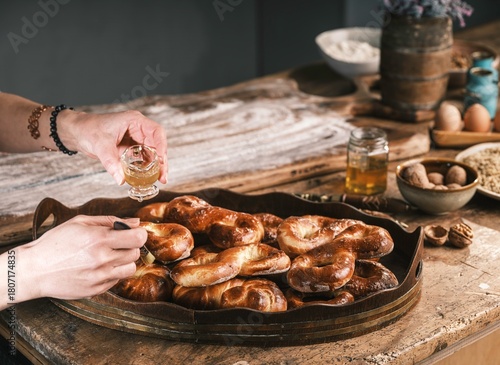 Hands are seen drizzling honey over freshly baked Romanian Mucenici pastries on a rustic wooden table