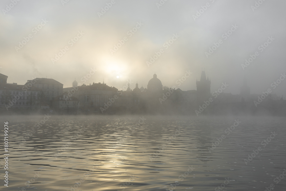 Naklejka premium Prague charles bridge appearing as silhouettes across the vltava river, bathed in the soft glow of a hazy sunrise