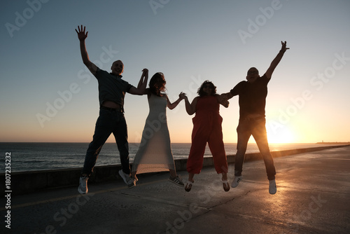 Four friends or family of four are jumping together at sunset near the ocean, holding hands and celebrating a joyful moment in silhouette.