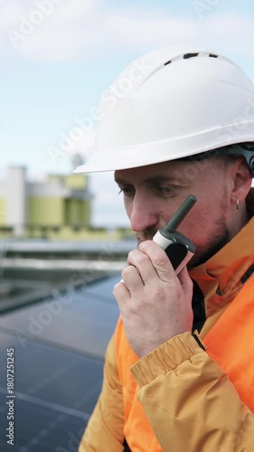 A solar panel installation engineer talks on a walkie-talkie on the roof of a building. Concept of clean energy and environmental sustainability. Vertical video.
