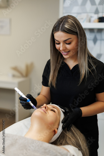 A beautician smiles while using a dermapen device for a microneedling treatment on a relaxed client lying on a spa bed, with a clean and modern clinic background
