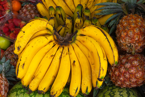 Ripe Juicy Yellow Bananas with Tropical Fruits in Background
A bunch of ripe, juicy yellow bananas with pineapples, strawberries, oranges and watermelon  in the background at a vibrant market in Kenya