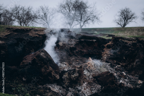 A rugged hillside emits white smoke from multiple cracks in the earth, creating a dramatic and surreal atmosphere.