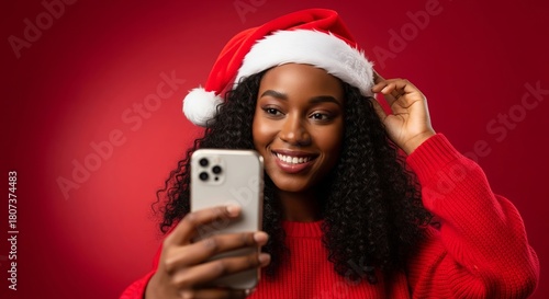 Joyful young African American woman in a festive Santa hat taking a happy selfie with her phone against a vibrant red background, celebrating the holiday season with delight and charm