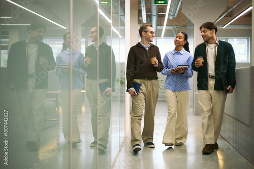 Three young adult colleagues, two Caucasian men and one Black woman, walking through modern office hallway holding coffee cups and digital tablet, engaging in conversation
